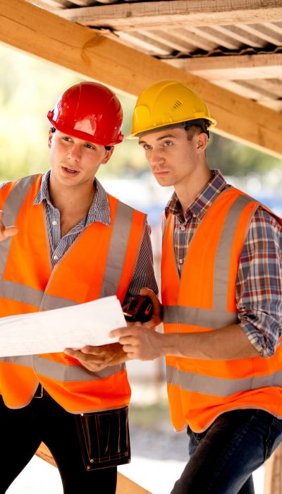 Two men dressed in shirts, orange work vests and helmets explore construction documentation on the building site near the wooden building constructions .